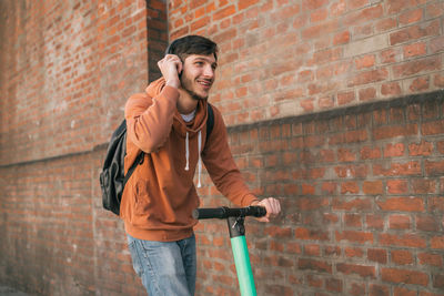 Young man listening music while riding push scooter against brick wall