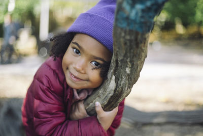 Portrait of smiling girl wearing knit hat and embracing tree branch at park