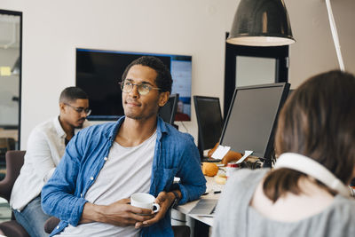 Thoughtful male computer programmer looking away while sitting by colleagues at desk in office