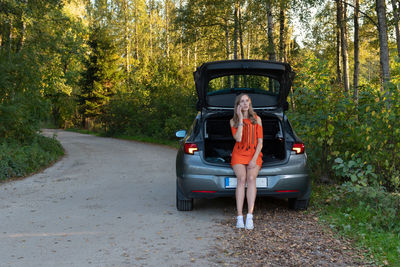 Woman talking on mobile phone while sitting on car against trees in forest