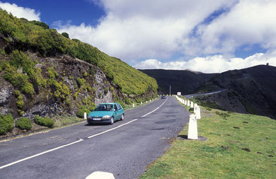 Car moving on road by mountain against cloudy sky