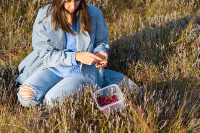 Young woman in denim trench sits on swamp sorting cranberries at autumn sunset