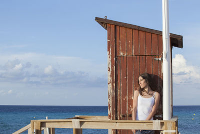 Smiling woman standing by lifeguard hut by sea against sky