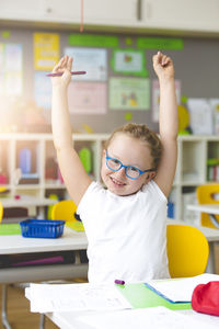 Portrait of girl with arms raised sitting at desk in classroom