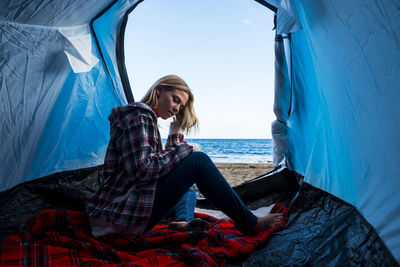 Woman sitting in tent at beach against sky