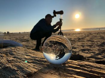 Reflection of man photographing at beach in crystal ball during sunset