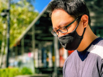 Young man in eyeglasses and face mask looking away against architecture, greenery and sky.