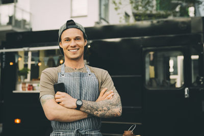 Portrait of smiling male owner with smart phone standing against commercial land vehicle