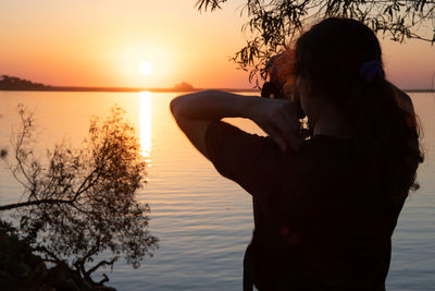 Rear view of woman standing by lake against sky during sunset