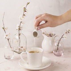 Woman pouring milk in stylish white cup of coffee among small vases with spring blossoming branches.
