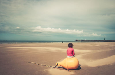 Rear view of girl sitting on buoy at beach against sky