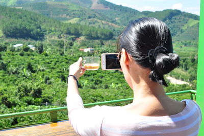 Rear view of woman photographing with mobile phone while holding coffee cup
