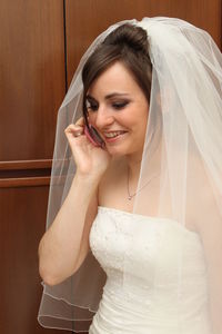 Smiling woman in wedding dress using phone while standing against wall