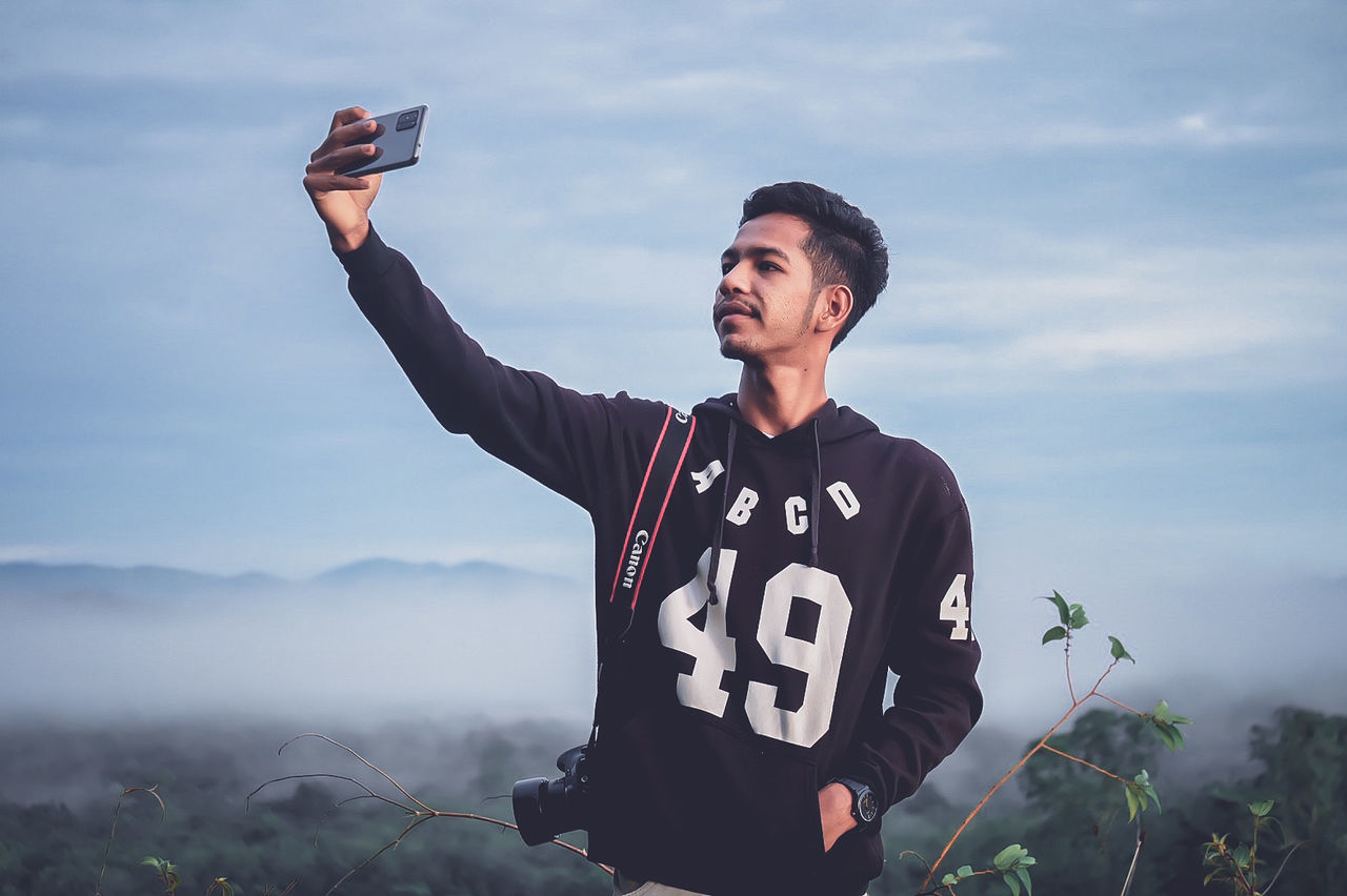 one person, technology, adult, communication, wireless technology, sky, mountain, activity, nature, sports, clothing, person, portable information device, smartphone, waist up, blue, outdoors, men, standing, emotion, mobile phone, smiling, camera, holding, portrait, happiness, lifestyles, tree, selfie, young adult, leisure activity, day, front view