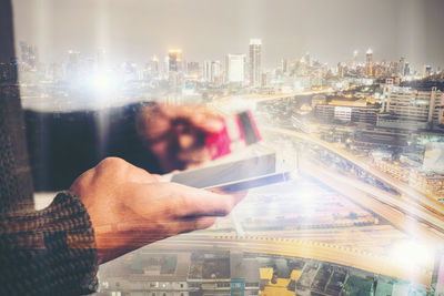 Multiple exposure of hands holding smart phone and card against illuminated modern buildings in city