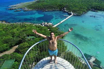 High angle portrait of shirtless man with arms outstretched at balcony against sea