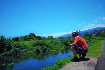 Side view of mid adult man crouching at riverbank against blue sky
