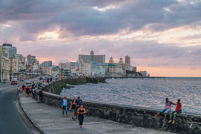 People in city by sea against sky during sunset