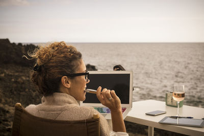 Rear view of woman looking away while sitting with laptop and alcohol at beach during sunset