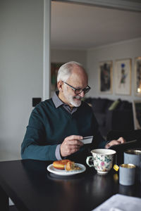 Smiling wrinkled man holding credit card while using digital tablet over table in living room