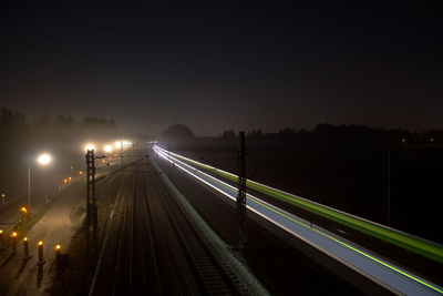 Light trails on road by railway tracks against sky at night