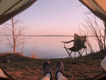 Low section of person by lake against sky during sunset