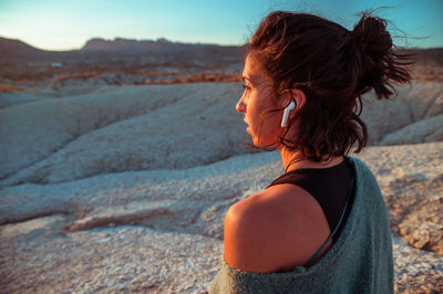 Side view of young female in sportive outfit and listening to music with wireless earbuds contemplating amazing scenery of rough stony badlands at sunset time