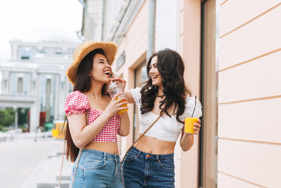 Happy young women friends in summer clothes with juice in hands having fun on summer city street