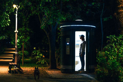 Man with french bulldog standing by public toilet in the park at night