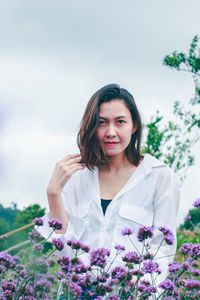 Portrait of a beautiful young woman standing by purple flowering plants against sky