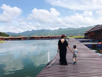Rear view of mother and son walking on footbridge over lake against cloudy sky