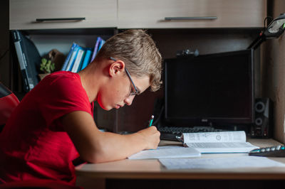 A teenage boy in a red t-shirt sits at a table at home and does his homework. study without gadgets.