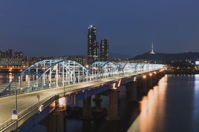 Illuminated bridge over river by buildings against sky at night