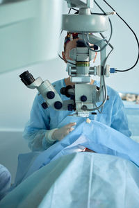 Adult female doctor in sterile mask and ornamental medical cap looking through surgical microscope against crop coworker in hospital