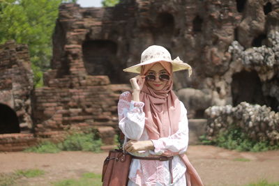 Portrait of woman wearing hat standing against stone wall