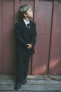 Thoughtful boy with hands clasped wearing suit while looking away against wooden wall