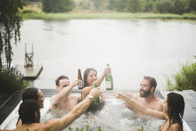 Carefree male and female friends enjoying drinks in hot tub against lake during weekend getaway