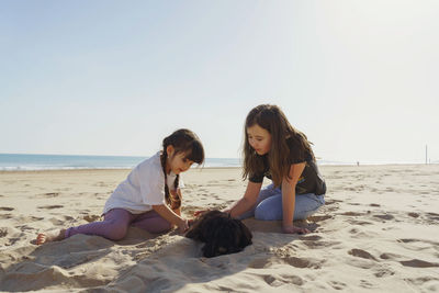 Side view of two girls sitting on beach and playing with dog