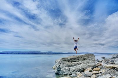 Boy jumping on rock against sky and lake