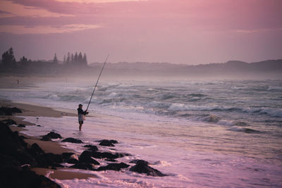 Man fishing in sea against sky during sunset