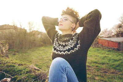 Young woman with hands behind head sitting on field against sky