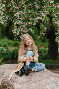 Caucasian cute baby girl with long wavy blonde hair in a trench coat sits on a stone in spring