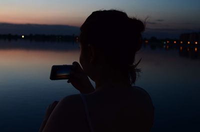Silhouette woman photographing lake against sky during sunset