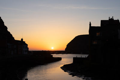 Silhouette buildings by sea against sky during sunset