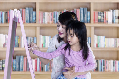 Mother assisting daughter in writing on blackboard while standing against bookshelf