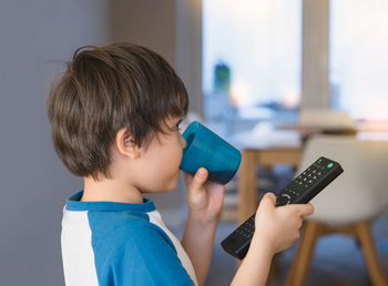 Side view of cute boy holding remote control drinking soda at home