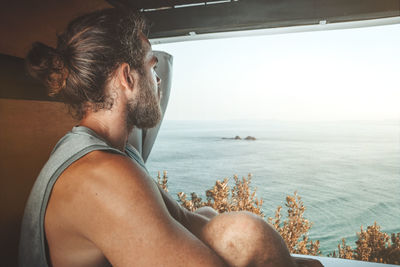 Young man sitting by sea against sky