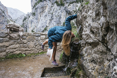 Full body of female hiker drinking cold fresh water from spring in rocky slope while trekking in mountainous terrain