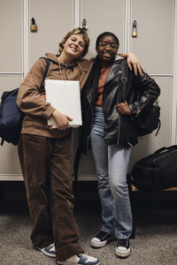 Full length portrait of smiling teenage girl standing with arm around female friend against locker in school