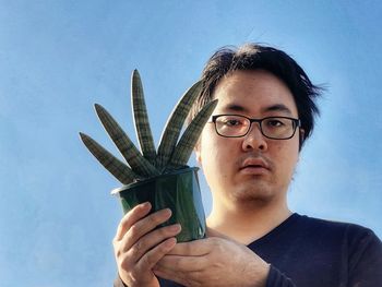 Portrait of young man holding eyeglasses against blue sky